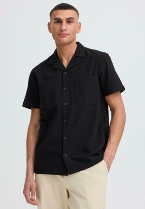 Giovane uomo con camicia nera a maniche corte con due tasche sul petto e pantaloni beige, in piedi con una mano in tasca.