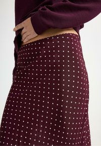 Burgundská sukně s puntíkovaným vzorem, měkkou strukturou, vlnkovaným pasem a přiléhavým střihem. Stylizováno s topem s dlouhým rukávem.