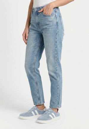 Person, die hellblaue Jeans mit geradem Bein und graue Sneaker mit weißen Streifen trägt, steht mit einer Hand in der Tasche.