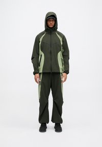Groene twee-delige buitenoutfit bestaande uit een jas met kap en contrasterende lichtgroene accenten, en bijpassende losse broek. Zwarte ritsen en schoenen.
