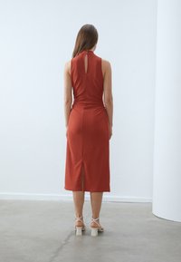 Midi šaty v rezavě červené barvě s texturovanou tkaninou, vysokým límcem, detaily ve tvaru klíčové dírky na zadní straně a bočním rozparkem. Nosené s světlými sandály na blokovém podpatku.