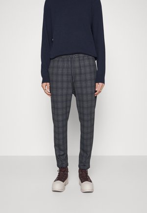 Grau karierte Hose mit einem Kordelzugbund, kombiniert mit einem navyblauen Langarmoberteil und beigen sowie braunen Schnürschuhen.