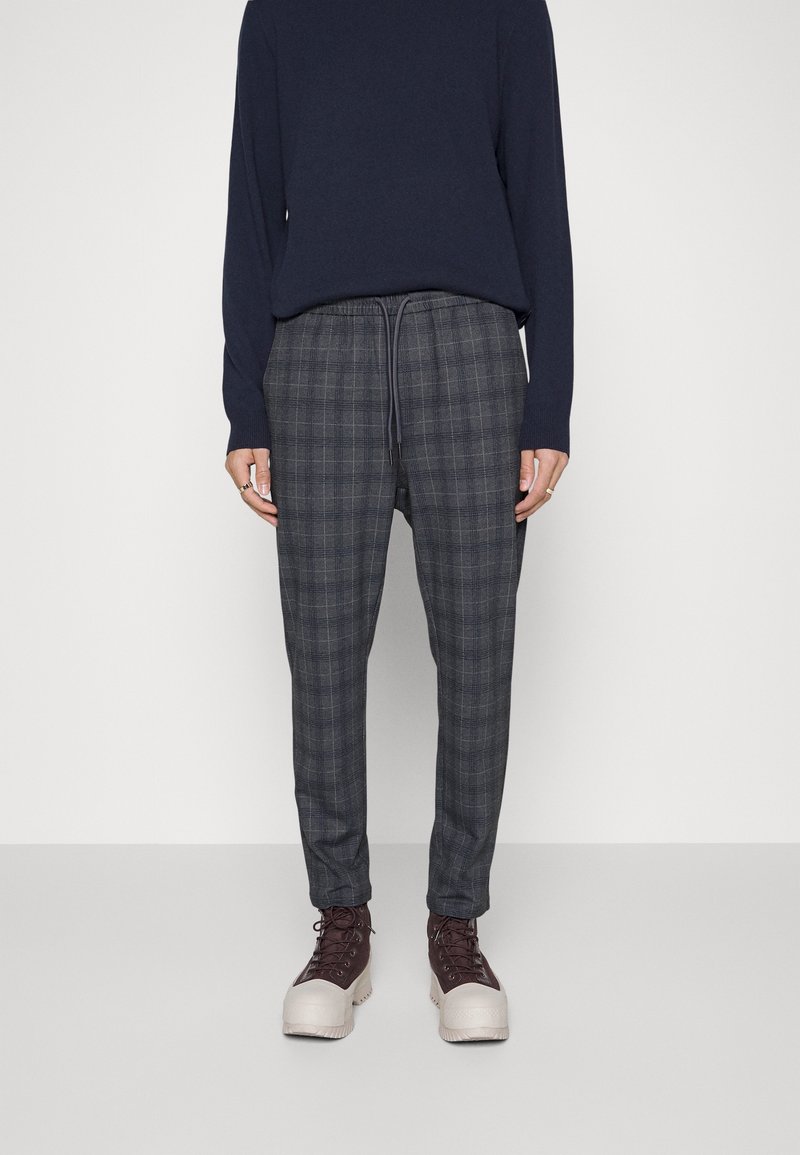 Grå rutiga byxor med dragsko i midjan, i kombination med en marinblå långärmad topp och beige och bruna snörkängor.