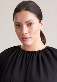 Czarny top z marszczonym dekoltem, gładką tkaniną, luźnym krojem i krótkimi rękawami. Tło jest neutralne i nie rzuca się w oczy.