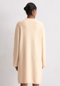 Robe-sweater longue et oversize en tissu beige doux, dotée d'un col arrondi, de longues manches et d'un ourlet droit. Texture lisse, détails minimaux.