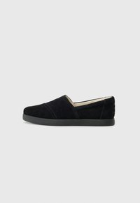 Scarpa slip-on in suede nera con suola in gomma bassa, caratterizzata da accenti di cuciture diagonali e una punta arrotondata.
