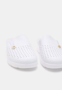 Vita slip-on skor med perforerat mönster, rund tå och en texturerad sula. Har en liten guldfinish på sidan.