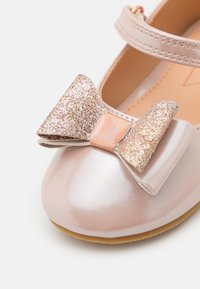 Zapato de cuero rosa pálido con un acabado brillante, que presenta un gran lazo de purpurina en color oro rosa y una puntera redondeada, asegurado por una correa.