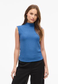 Blå ärmlös topp med hög halsringning, texturerad tyg, och en figurformad design, tillsammans med svarta byxor. Knappar syns inte.