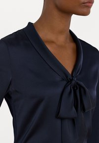 Donkerblauwe blouse met een zachte, satijnen textuur. Beschikt over een sjawlcollar en een strik bij de halslijn, die een soepele, elegante uitstraling benadrukt.