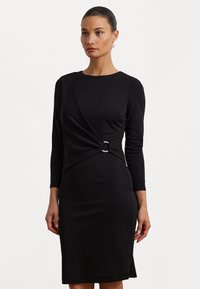 Zwarte jurk met lange mouwen, voorzien van een gedraaide design bij de taille en een metalen ringaccent. Glad, rekbaar stof.