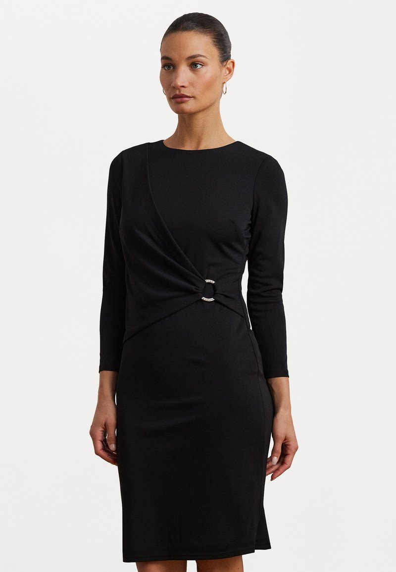 Zwarte jurk met lange mouwen, voorzien van een gedraaide design bij de taille en een metalen ringaccent. Glad, rekbaar stof.