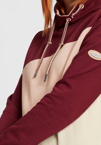 Femme portant un sweat à capuche bicolore avec des manches bordeaux, des sections beige et rose, des cordons de serrage, et un patch logo Oximo sur le bras gauche.