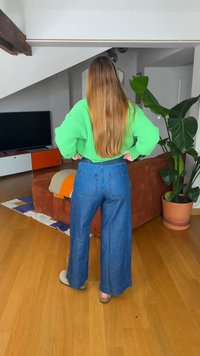 Γυναίκα με μακριά μαλλιά που φοράει ένα φωτεινό πράσινο πουλόβερ και φαρδιά μπλε τζιν, στέκεται με τα χέρια στη μέση σε ένα σαλόνι με ένα μεγάλο φυτό.