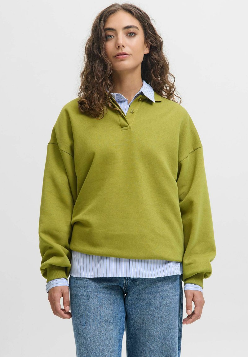 Woman with curly brown hair wearing a green pullover over a blue striped shirt and blue jeans, standing against a plain background.