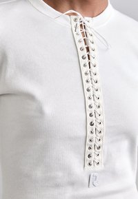 Wit geribbelde shirt met lange mouwen, voorzien van een voorpaneel met vetersluiting met zilveren oogjes en een klein logo aan de onderzijde links.