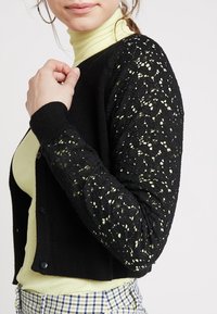 Svart kofta med spetsärmar, med ett blommönster. Kroppen är ribbad med knappar, och den är kombinerad med en ljusgrön polotröja under.