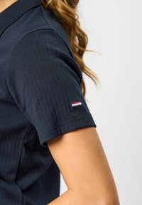 Marineblå ribbestrikket kortermet skjorte på person med lys hud og langt bølget brunt hår, liten rød, hvit og blå lapp på ermet.