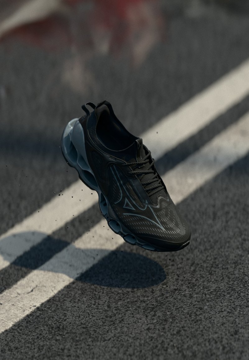 Zapatilla deportiva negra con parte superior texturizada, que presenta un diseño elegante y una suela translúcida acolchada, mostrada en el aire sobre una superficie de carreteras.
