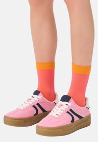 Rosafarbene Wildledersneaker mit einem marineblauen Design an der Seite und cremefarbenen Schnürsenkeln, kombiniert mit lebhaft pinken und orangefarbenen gerippten Socken.