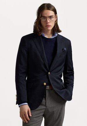 Blazer marine avec boutons dorés, porté sur un pull marine et une chemise bleu clair. Un pantalon gris et un mouchoir de poche à pois complètent le look.