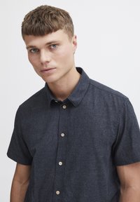 Camisa de manga corta, de color azul oscuro, hecha de tejido texturizado, con cuello, cierre delantero de botones de madera y un ajuste relajado.