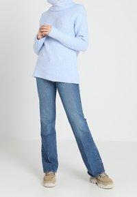 Kvinna i ljusblå polo-tröja, raka blå jeans och beige sneakers som står med händerna i knä.