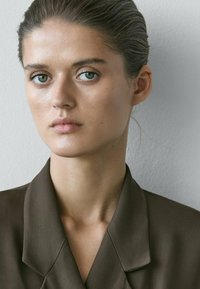 Junge Frau mit zurückgebundenem Haar, die einen dunkelbraunen Blazer trägt und direkt nach vorne blickt, vor einem einfarbigen, hellen Wandhintergrund.