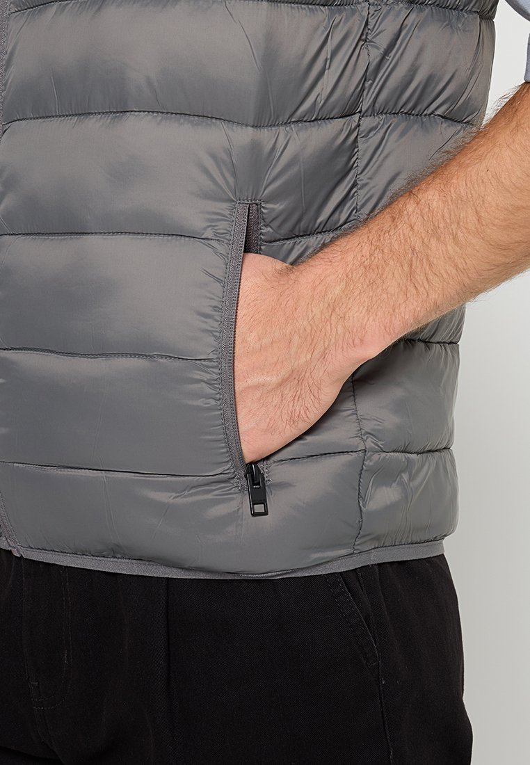 Chaleco acolchado gris con patrón de rombos horizontal, bolsillo lateral con cremallera, textura suave y escote redondeado. Se llevan pantalones negros debajo.