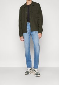 Olivgrön ulljacka med knappstängning och ficka, bärs över en svart skjorta, ihopparad med ljusblå jeans och flerfärgade sneakers.