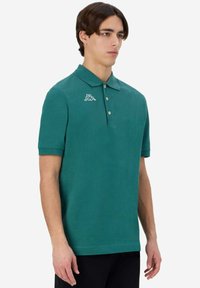 Polo verde realizzato in un tessuto morbido, con colletto classico, patta con tre bottoni e un logo bianco sul lato sinistro del petto.