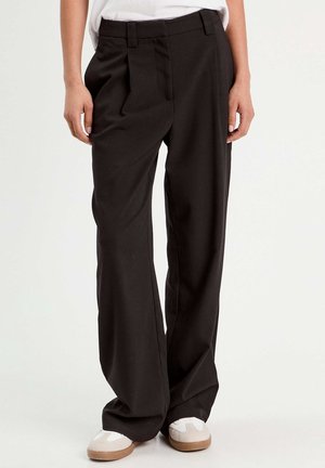 Personne portant un pantalon noir ample et des baskets blanches, debout devant un fond clair uni.