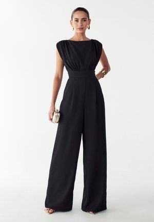 Black wide-leg jumpsuit with a boat neckline, pleated shoulders, and a cinched waist. Model holds a gold clutch and wears gold accessories.