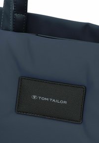Sac fourre-tout navy avec un patch en tissu noir texturé arborant le logo "TOM TAILOR". Matériau lisse, poignées robustes, design simple.