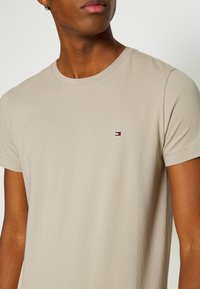 Beige bomulds t-shirt med kortærmer, rund halsudskæring og lille broderet logo med røde, hvide og blå accenter på brystet.