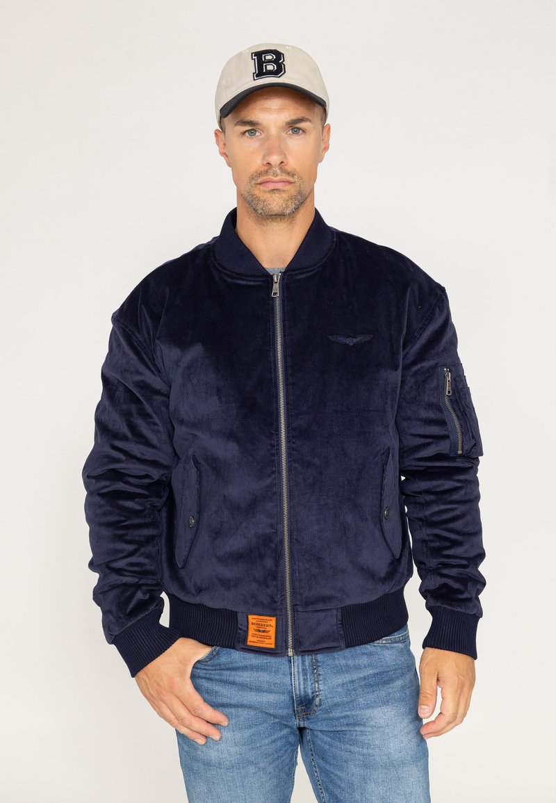 Veste bomber bleu marine en matériau doux et velouté avec des poignets côtelés, deux poches avant et une poche à fermeture éclair sur la manche. Étiquette orange au bas.