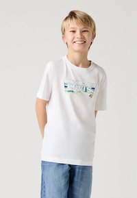 Weißes Baumwoll-T-Shirt mit einem farbenfrohen Levi's-Logo. Kombiniert mit hellblauen Jeans. Kurze Ärmel, Rundhalsausschnitt.