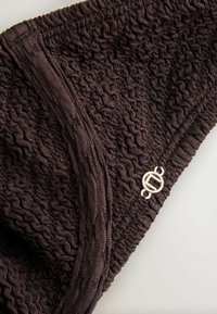 Țesătură tricotată maro închis cu model ondulat, având o cusătură verticală și o mică clemă rotundă metalică pe un fundal deschis.