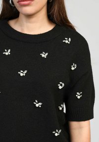 Pull en tricot noir avec un col rond, orné de perles et de cristaux en floral, répartis uniformément sur le devant. Manches courtes.