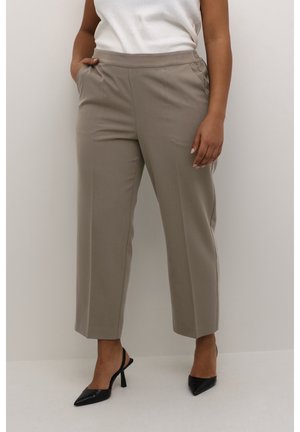 Kobieta nosząca taupe spodnie o wysokim stanie, dopasowane, z bocznymi kieszeniami, w zestawieniu z czarnymi szpilkami typu slingback z ostrym noskiem oraz białym bezrękawnikiem.