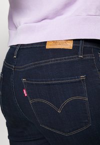 Jeans de mezclilla azul oscuro con una etiqueta de cuero en la cinturilla trasera. Presentan costuras amarillas y un diseño de bolsillo distintivo en forma de corazón.