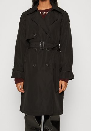 Frau trägt einen schwarzen, zweireihigen Trenchcoat mit Gürtel über einem dunkelroten Pullover und glänzenden schwarzen Hosen, steht vor einem schlichten Hintergrund.