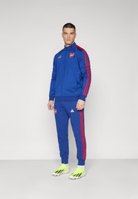 Männlicher Sportanzug in königsblau mit roten Streifen, mit einem Arsenal-Logo, aus glattem Material, kombiniert mit leuchtend grünen Fußballschuhen.