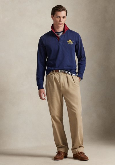 Μπλε σκούρο πόλο με μακριά μανίκια και κόκκινο γιακά, με χρυσό έμβλημα λιονταριού, συνδυασμένο με μπεζ παντελόνια και καφέ παπούτσια, στυλιζαρισμένο με ριγωτό πουκάμισο από μέσα.
