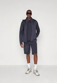 Ciemnoszara sportowa kurtka z dopasowanymi szortami, oba elementy posiadają zamki błyskawiczne. Model ma na sobie okulary przeciwsłoneczne i trampki, stoi na neutralnym tle.