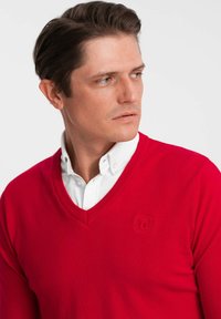 Czerwony sweter z dekoltem w serek, wykonany z teksturowanej dzianiny, z haftowanym logo na piersi, nałożony na białą koszulę z kołnierzykiem.