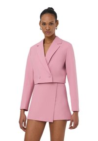 Blazer rosa accorciato con design doppiopetto, rever, e texture liscia, abbinato a una mini gonna coordinata con uno spacco laterale.