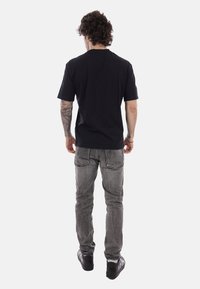 Uomo con capelli ricci, tatuaggi sul braccio sinistro, indossa una maglietta nera, jeans grigi e sneakers nere, in piedi di spalle su sfondo bianco.