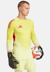 Geel doelmanstrui met rode accenten, lange mouwen, gestructureerd patroon. Zwarte handschoenen, houdt een voetbal vast met oranje en grijze details.
