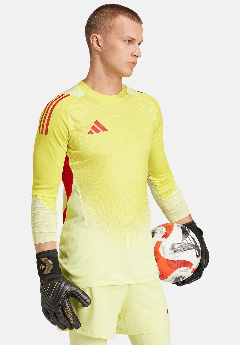 Geel doelmanstrui met rode accenten, lange mouwen, gestructureerd patroon. Zwarte handschoenen, houdt een voetbal vast met oranje en grijze details.